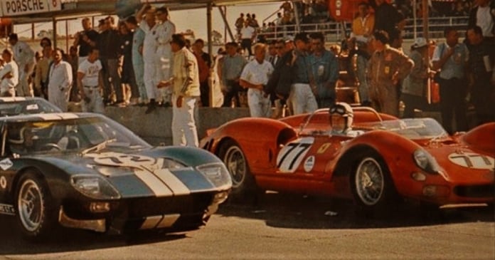 A black Ford GT40 and a red Ferrari race car, both with racing numbers, are positioned side by side at a pit stop with crews and spectators standing nearby.